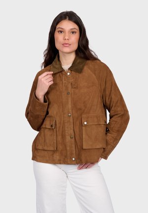 Frau trägt eine braune Wildlederjacke mit Klappentaschen und Knopfdetails, kombiniert mit weißen Hosen, und posiert vor einem schlichten Hintergrund.