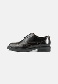 Sorte skinnsko med brogue-detaljer, slank avrundet form, svart såle og kontrasterende oransje sømmer langs sømmene.