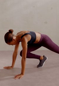 Fekete sportmelltartó és bordó leggings egy dinamikus edzési pozícióban lévő személyen, szürke sportcipőben egy semleges színű padlón.