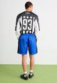 Adidas dres v čierno-bielych prúžkoch s číslom 93 na chrbte, v kombinácii s modrými šortkami a čiernymi topánkami, na zelenej podlahe.
