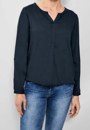 Mujer con una blusa negra de manga larga con un pequeño escote en V y vaqueros azules, de pie frente a un fondo liso y claro.