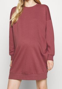 Abito oversize a maniche lunghe in un tessuto rosso-marrone tenue. Collo rotondo e polsini a costine, dotato di tasche laterali e una vestibilità rilassata.