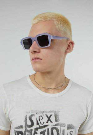 Lila zonnebril met een dikke, hoekige frame en donkere lenzen. Model draagt een wit T-shirt met zwarte tekst en een dunne zilveren ketting.