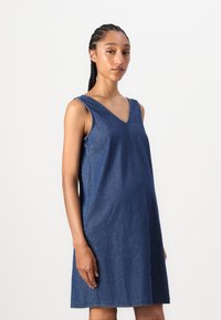 Vestido de ganga sem mangas com decote em V, ajuste descontraído e comprimento até ao joelho, na cor azul escuro com detalhes de costura visíveis.