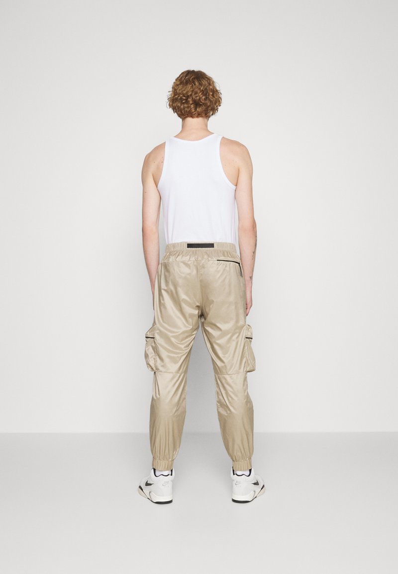 Pantalon cargo beige en matériau brillant, avec des poignets élastiques, des poches latérales et une taille avec cordon de serrage. Porté avec un débardeur blanc.
