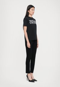 Juodas medvilninis marškinėlis su baltu "VERSACE JEANS COUTURE" logotipu, derinamas su juodomis siaurėjančiomis kelnėmis ir smailėmis juodomis aukštakulnėmis.