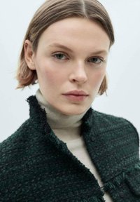 Zeleno teksturirana tweed jakna s raztrganimi robovi, preložena čez bež turtleneck. Poudarjen bližnji posnetek ovratnika in podrobnosti blaga.