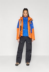 Μονωμένο πορτοκαλί και μπλε μπουφάν με γκρι λεπτομέρειες, φορεμένο πάνω από μπλε φλις μπλούζα, συνδυασμένο με μαύρο αδιάβροχο παντελόνι και υπαίθριες μπότες.