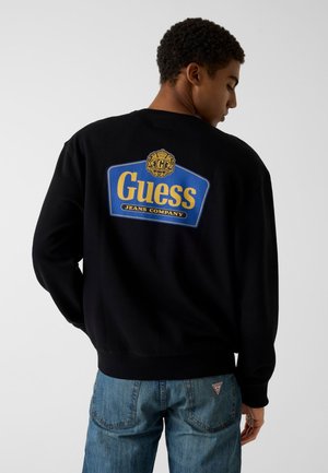 Joven de espaldas que lleva una sudadera negra con el logo azul y amarillo de "Guess Jeans Company" y vaqueros azules con un pequeño parche rojo en forma de triángulo.