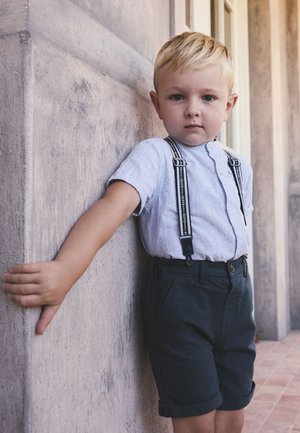 Ung dreng med blondt hår iført en lys skjorte med seler og mørke shorts læner sig op ad en tekstureret væg i en udendørs korridor.