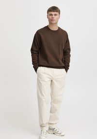 Brun genser med lange ermer, rund hals; kombinert med kremfargede, plisserte bukser og hvite joggesko med blå detaljer.