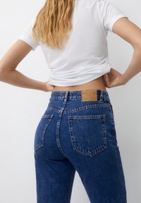 Denim jeans in dark blue with a high-waisted design, featuring two back pockets and a brown label at the waistband. White cropped t-shirt.