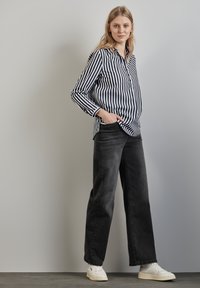 Camicia a righe bianco e nere con bottoni e maniche lunghe, abbinata a jeans neri oversize e sneakers bianche su uno sfondo neutro.