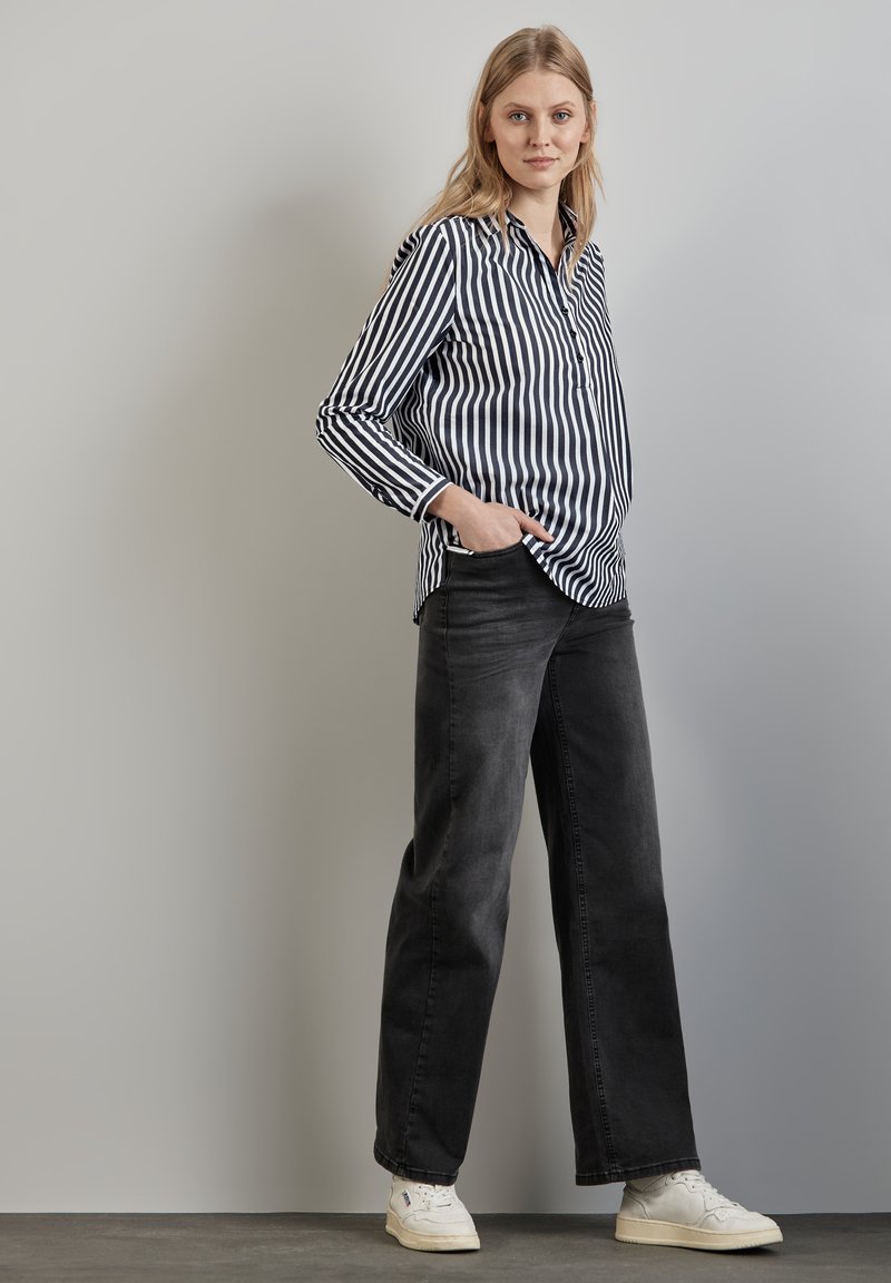 Camicia a righe bianco e nere con bottoni e maniche lunghe, abbinata a jeans neri oversize e sneakers bianche su uno sfondo neutro.