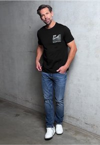 Camiseta de algodón negra con un estampado gráfico en el bolsillo, combinada con jeans de denim azules y zapatillas blancas. De pie contra una pared de hormigón.