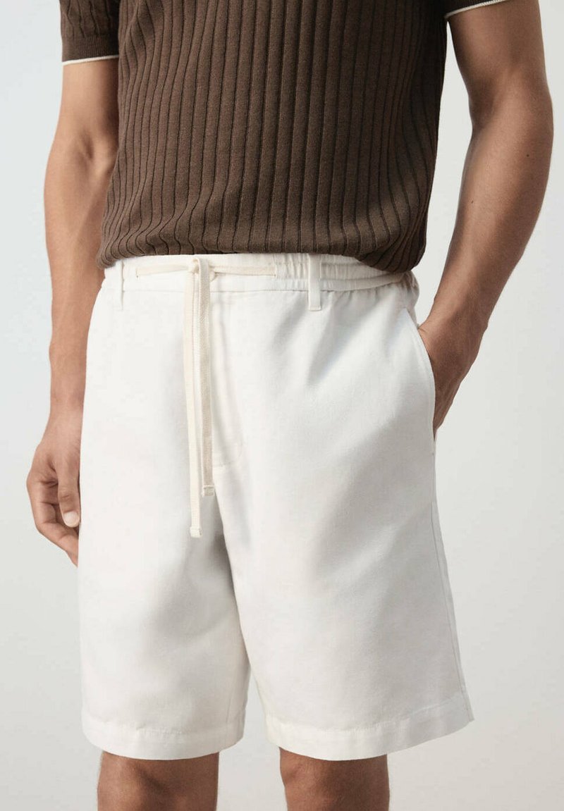 Homme portant un short blanc à cordon et une chemise marron côtelée à manches courtes, une main dans la poche sur fond clair.