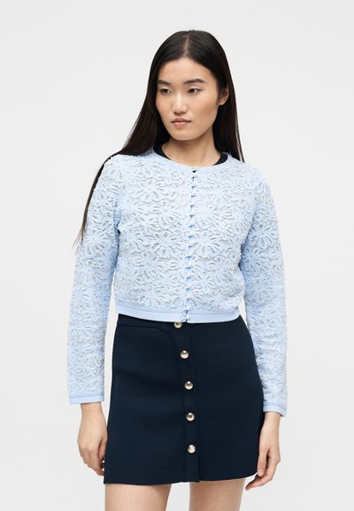 Νεαρή γυναίκα που φοράει ανοιχτό γαλάζιο δαντελένιο ζακέτα με κουμπιά από πέρλες και φούστα με κουμπιά μπροστά σε ναυτικό μπλε χρώμα, με φόντο απλό.