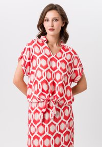 Blouse à motifs rouge et blanc avec un col en V, des manches courtes et un lien à la taille, fabriquée en tissu léger. Design géométrique hexagonal.