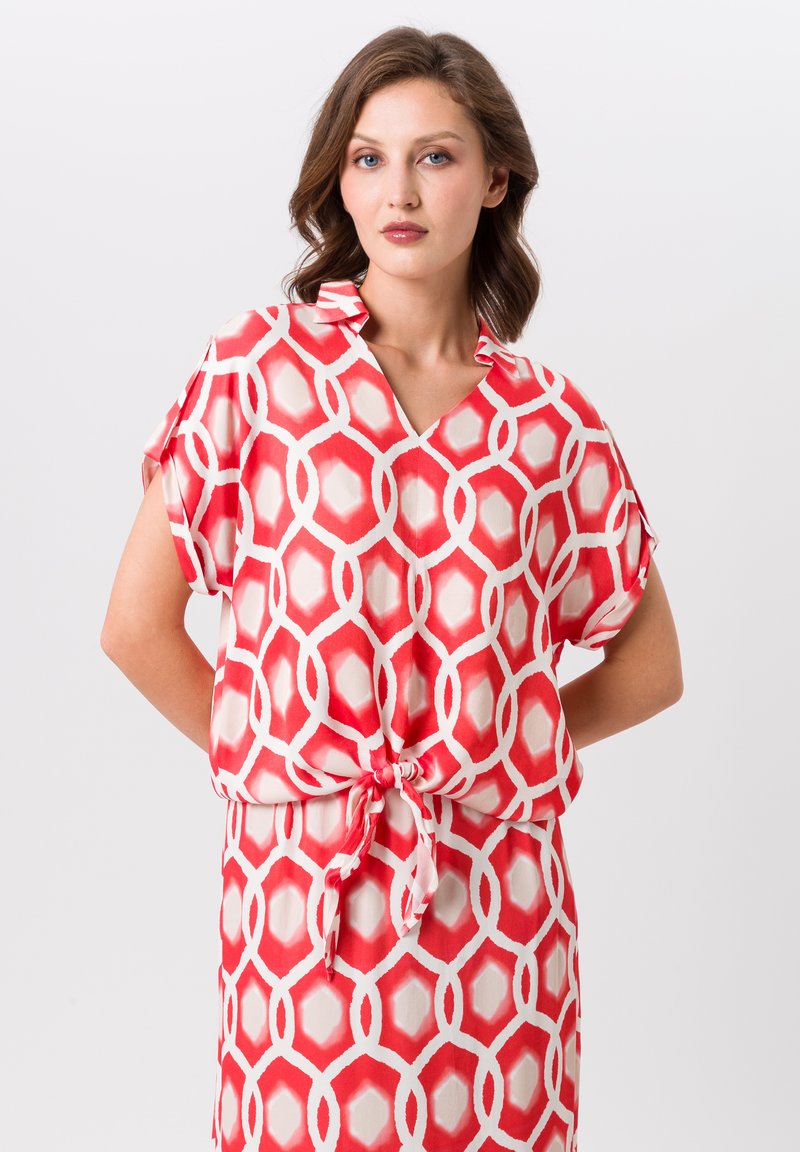 Blouse à motifs rouge et blanc avec un col en V, des manches courtes et un lien à la taille, fabriquée en tissu léger. Design géométrique hexagonal.