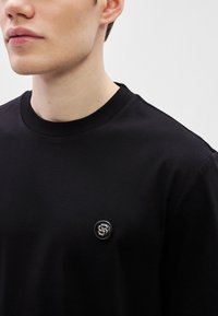 Μαύρο βαμβακερό t-shirt με κοντά μανίκια, με στρογγυλή λαιμόκοψη και μικρό, κυκλικό λογότυπο στο στήθος σε μαύρο και ασημί.