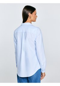 Sieviete ar tumšiem matiem valkā gaisā zilu kreklu ar pogām un zilas džinsas, redzama no muguras un sānskata vienkārša fona priekšā.