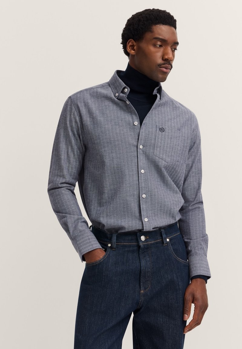 Uomo che indossa una camicia grigia con bottoni sopra un dolcevita nero e jeans scuri blu, in piedi con una mano in tasca, mentre guarda di lato.