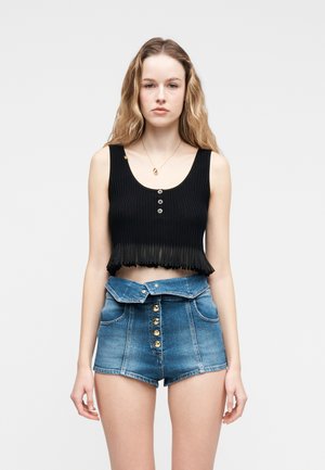 Młoda kobieta o falowanych blond włosach, ubrana w czarny, bezrękawnikowy top typu crop oraz wysokiej talii niebieskie dżinsowe szorty z złotymi guzikami.