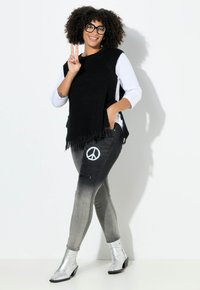 Poncho de punto negro con flecos, superpuesto a una camisa blanca. Jeans grises con un estampado de símbolo de paz, combinados con botas de tobillo plateadas.