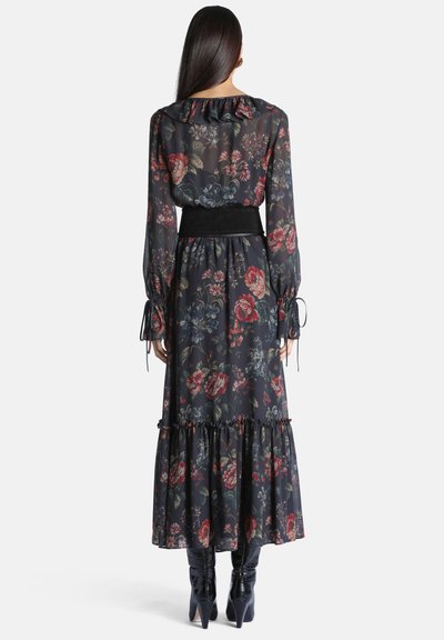 Femme aux longs cheveux foncés portant une robe maxi noire à fleurs à manches longues avec des volants et des bottes à talons noires, debout de dos.
