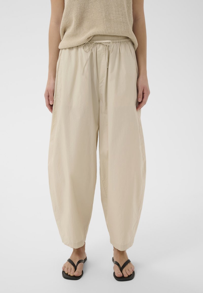 Persona vestida con pantalones beige sueltos con cordón y un top de punto sin mangas beige, de pie con las manos a los lados y chanclas negras.