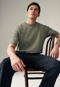 Kortærmet olivengrøn bomuld t-shirt, afslappet pasform, kombineret med mørke teksturerede bukser, siddende på en træstol foran en neutral baggrund.