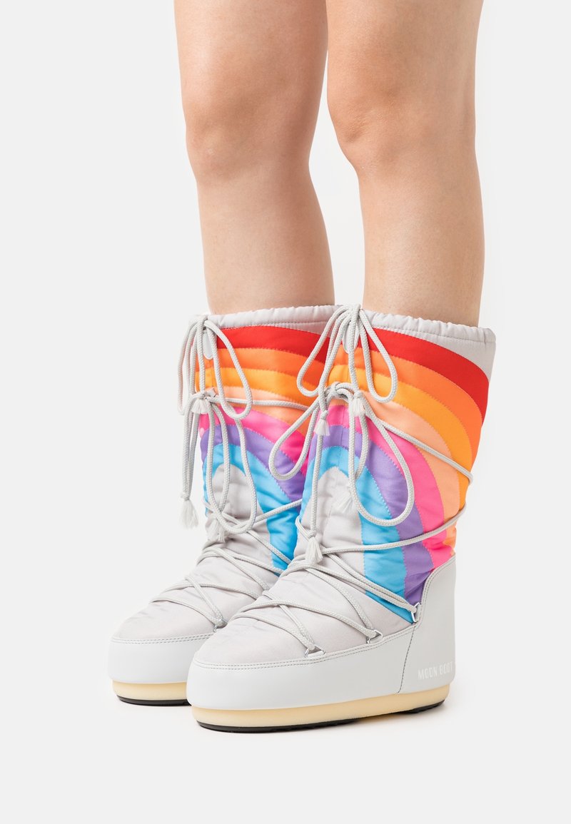 Jambes portant des bottes d'hiver blanches matelassées avec des rayures arc-en-ciel et des lacets gris clair noués autour des mollets sur un fond blanc.