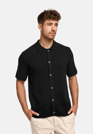 Man with curly hair wearing a black short-sleeve button-up knit shirt and light beige pants, hands in pockets, looking right.