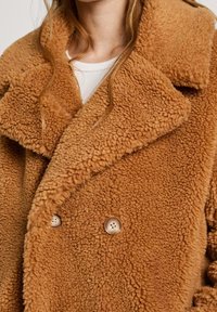 Brązowy, puszysty płaszcz dwurzędowy wykonany z fakturowanej tkaniny, z dużymi wyciętymi klapami i okrągłymi guzikami z czterema otworami.