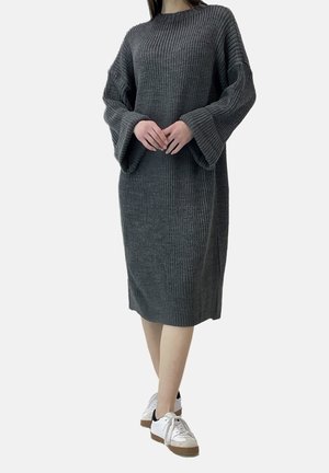Personne portant une robe-pull en maille côtelée longue gris foncé avec de larges manches et des baskets blanches, debout, les mains légèrement jointes.