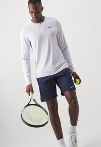 Camisa atlética blanca de manga larga con cuello de cremallera, combinada con pantalones cortos azul marino. Sosteniendo una raqueta de tenis y una pelota, con zapatillas blancas.