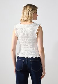 Hvit smocket topp med volangermer, teksturert overflate og rynket kant, kombinert med mørkeblå denimjeans med baklommer.