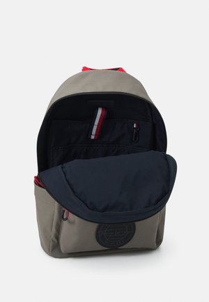 Sac à dos beige et noir avec poche avant et languettes à rayures rouge, blanc et bleu, ouvert pour révéler les compartiments intérieurs.