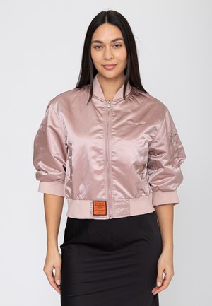 Donna con capelli lunghi e scuri che indossa un bomber rosa chiaro lucido con cerniera e una gonna nera, in piedi davanti a uno sfondo neutro.