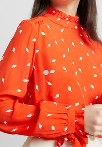 Vrouw in een heldere oranje blouse met witte bladpatronen, gerimpelde kraag en gestrikte manchetten, weergegeven van nek tot taille.