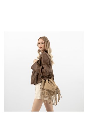 Jeune femme aux cheveux longs blonds portant une veste en cuir marron et un short beige, avec un sac beige à franges sur l'épaule.