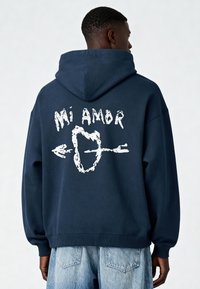 Námornícka mikina s kapucňou a bielym grafickým textom "Mi Amor" a dizajnom šípu na chrbte. Vyrobená z príjemnej látky s rebrovanými manžetami a lemom.