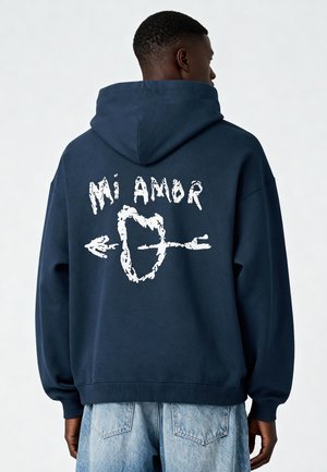 Tumši zils kapuce krekls ar baltu grafiku "Mi Amor" un bultas dizainu uz muguras. Izgatavots no mīksta auduma ar ribinātu piedurkņu un apakšmalu.