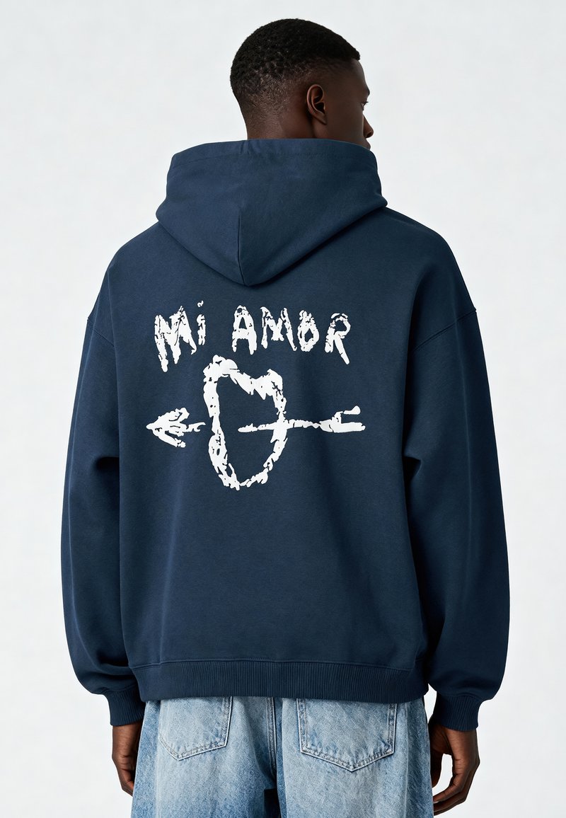 Námornícka mikina s kapucňou a bielym grafickým textom "Mi Amor" a dizajnom šípu na chrbte. Vyrobená z príjemnej látky s rebrovanými manžetami a lemom.