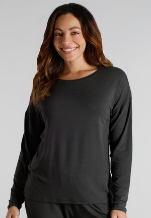 Smiling woman with wavy brown hair wearing a loose black long-sleeve shirt against a plain light gray background.
