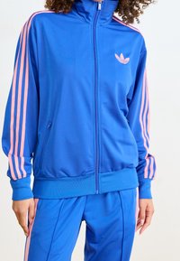 Blauwe trainingspak met roze strepen op de mouwen en broek, met ritsjack, en het Adidas-logo op de borst, gedragen door een persoon met krullend haar.