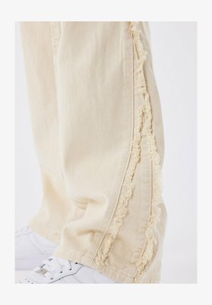 Un pantalon beige en denim avec des coutures latérales frangées et une coupe décontractée, associé à des baskets blanches, visibles au bas.