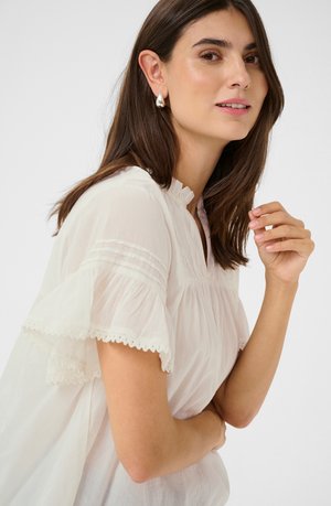 Witte, lichte blouse met ruches aan de mouwen, een gegatherde halslijn en decoratieve kantafwerking langs de randen. Soepele, zachte textuur.