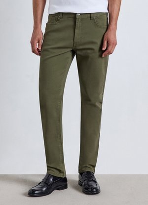 Uomo che indossa pantaloni verde oliva a gamba dritta, mocassini in pelle neri e una camicia bianca, in piedi su un pavimento grigio.