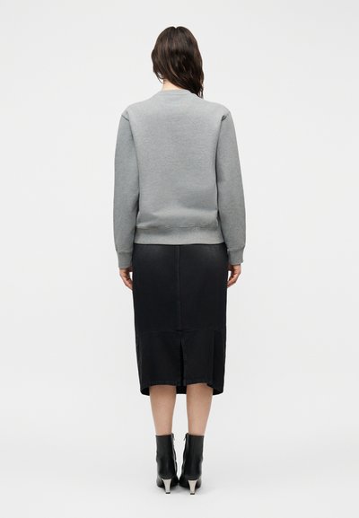 Frau mit langen dunklen Haaren, die einen grauen Pullover, einen schwarzen Midirock mit Rücken-Schlitz und schwarze Stiefeletten mit silbernen Absätzen trägt, steht mit dem Rücken zur Kamera.